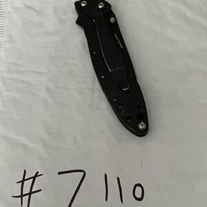 May include: A black folding knife with a pocket clip, visible screws, and the text "489PVTCT". The number "7110" is handwritten in black ink below the knife on a white surface.