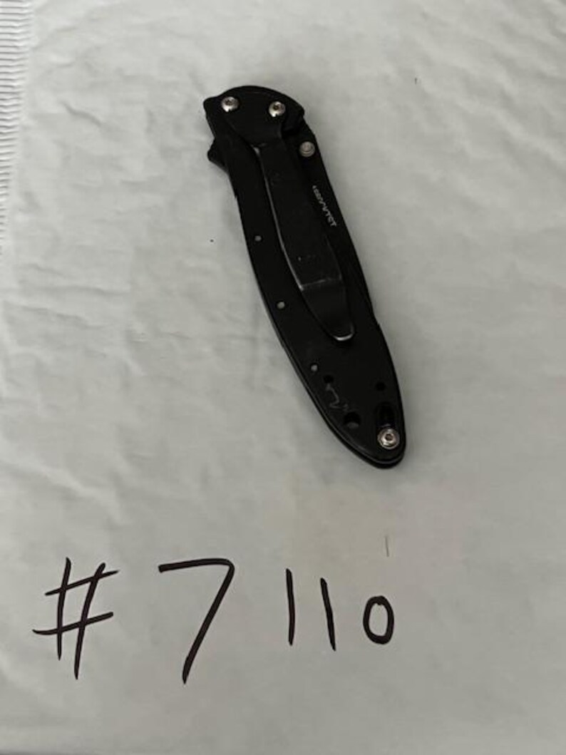 May include: A black folding knife with a pocket clip, visible screws, and the text "489PVTCT". The number "7110" is handwritten in black ink below the knife on a white surface.