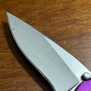 May include: Close-up of a silver folding knife blade with a purple handle. The blade is sharp and has a slight curve. The knife is partially open, revealing the blade's edge. The handle is a vibrant purple color.
