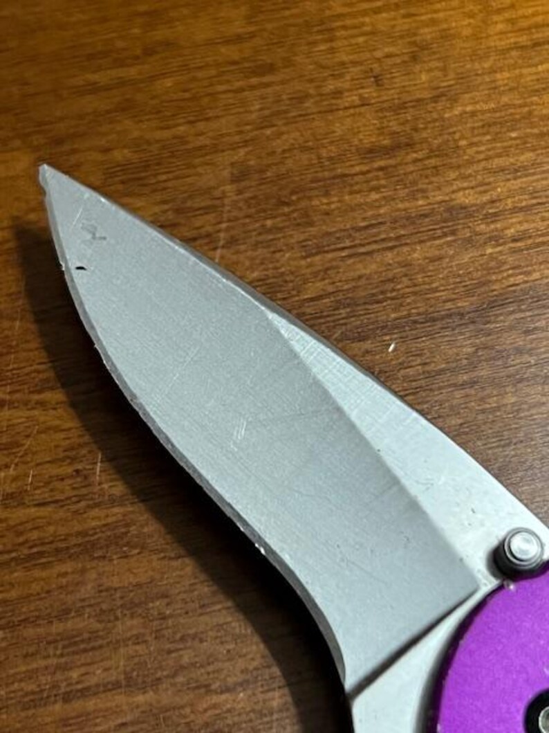 May include: Close-up of a silver folding knife blade with a purple handle. The blade is sharp and has a slight curve. The knife is partially open, revealing the blade's edge. The handle is a vibrant purple color.