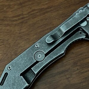 Puede incluir: Una navaja plegable con un acabado met&aacute;lico gris oscuro desgastado en el mango. El mango cuenta con un clip de bolsillo de metal, cabezas de tornillos visibles y una secci&oacute;n recortada. La superficie del metal tiene un aspecto texturizado y mate.