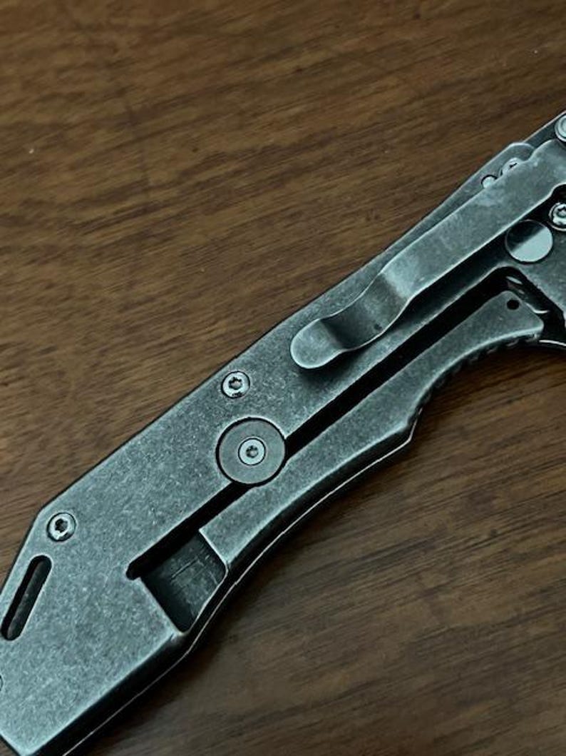 Puede incluir: Una navaja plegable con un acabado met&aacute;lico gris oscuro desgastado en el mango. El mango cuenta con un clip de bolsillo de metal, cabezas de tornillos visibles y una secci&oacute;n recortada. La superficie del metal tiene un aspecto texturizado y mate.