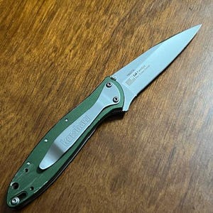 May include: A green and silver folding knife with a silver blade. The knife has a pocket clip with the brand name "Kershaw" printed on it. The blade has text that includes "1660OL" and "Kai Onion Design".