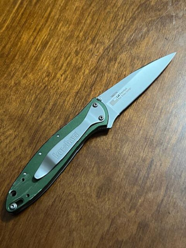 May include: A green and silver folding knife with a silver blade. The knife has a pocket clip with the brand name "Kershaw" printed on it. The blade has text that includes "1660OL" and "Kai Onion Design".