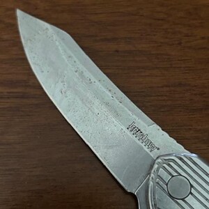 May include: Close-up of a Kershaw folding knife with a silver blade and handle. The blade shows signs of wear. The handle has a textured design and a circular button. The knife is on a brown wooden surface.