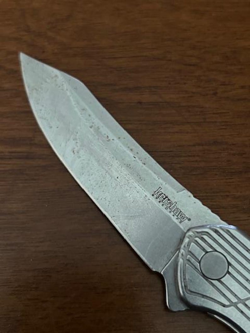 May include: Close-up of a Kershaw folding knife with a silver blade and handle. The blade shows signs of wear. The handle has a textured design and a circular button. The knife is on a brown wooden surface.