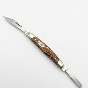 May include: A folding pocket knife with two silver-toned metal blades and a brown wood-grain handle. The handle features a silver oval inlay with the text BUCK. One blade is stamped with the text BUCK 555. The knife is shown in an open position.