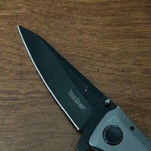 K&ouml;nnte beinhalten: Nahaufnahme eines Kershaw-Klappmessers mit schwarzer Klinge und grauem Griff. Der Markenname "Kershaw" ist auf der Klinge aufgedruckt. Das Messer liegt auf einer Holzoberfl&auml;che.