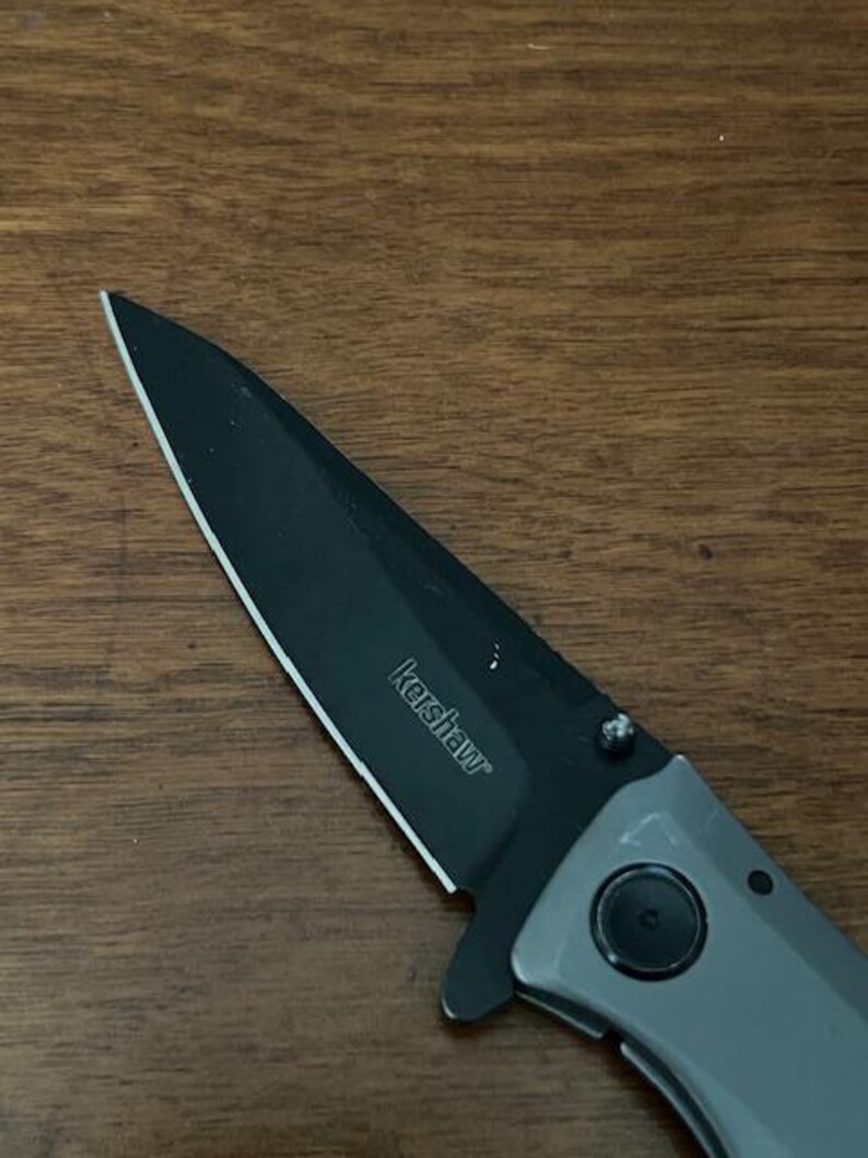 K&ouml;nnte beinhalten: Nahaufnahme eines Kershaw-Klappmessers mit schwarzer Klinge und grauem Griff. Der Markenname "Kershaw" ist auf der Klinge aufgedruckt. Das Messer liegt auf einer Holzoberfl&auml;che.