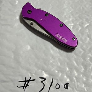 May include: A purple Kershaw folding knife with a silver blade. The knife has a textured handle and is closed. The text "Kershaw" is visible on the side of the knife. The number "#3100" is written below the knife.