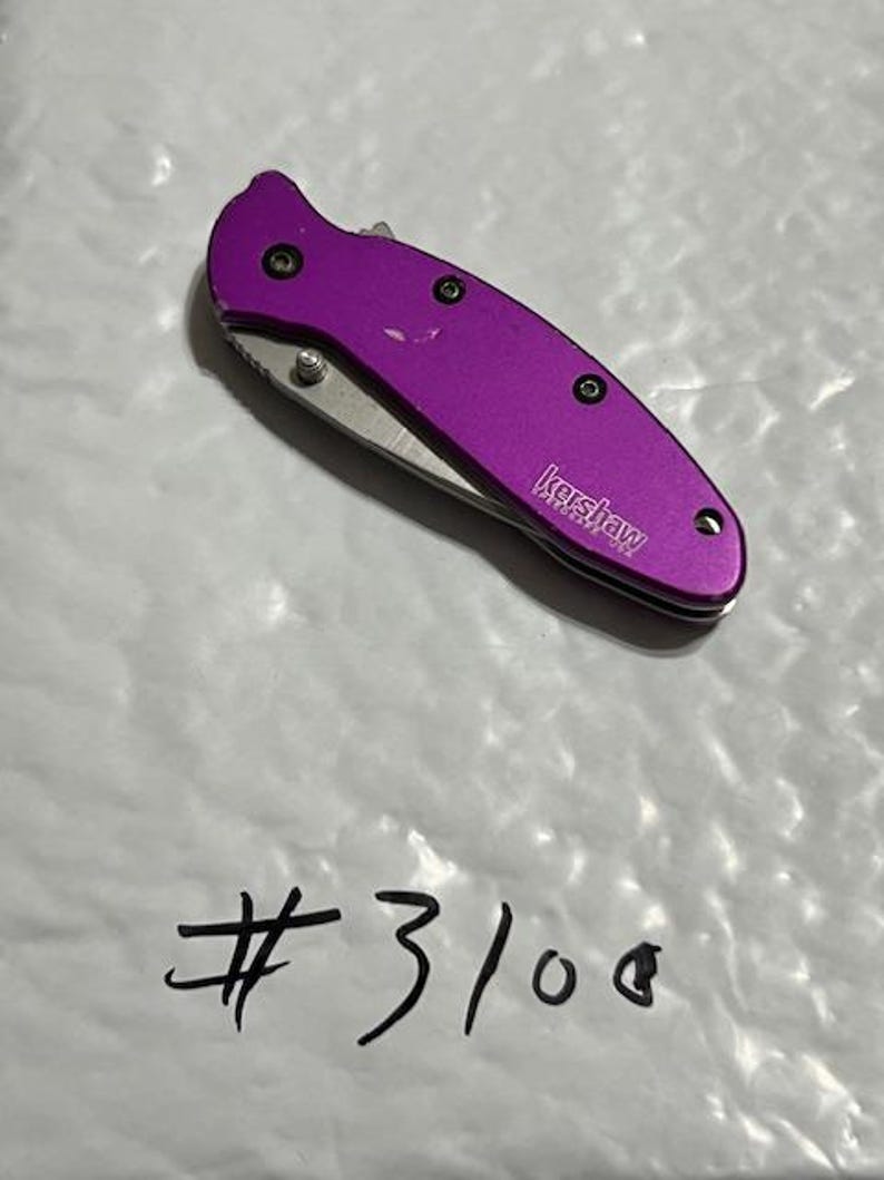 May include: A purple Kershaw folding knife with a silver blade. The knife has a textured handle and is closed. The text "Kershaw" is visible on the side of the knife. The number "#3100" is written below the knife.