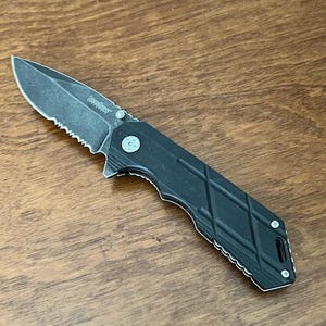 May include: A black folding knife with a partially serrated blade. The knife has a textured handle with a diamond pattern. The blade is dark gray and the knife is lying on a wooden surface.