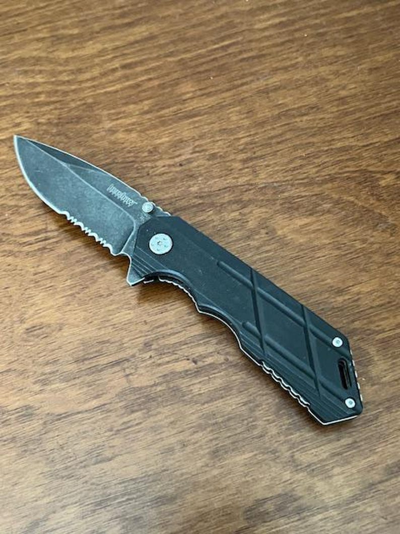 May include: A black folding knife with a partially serrated blade. The knife has a textured handle with a diamond pattern. The blade is dark gray and the knife is lying on a wooden surface.