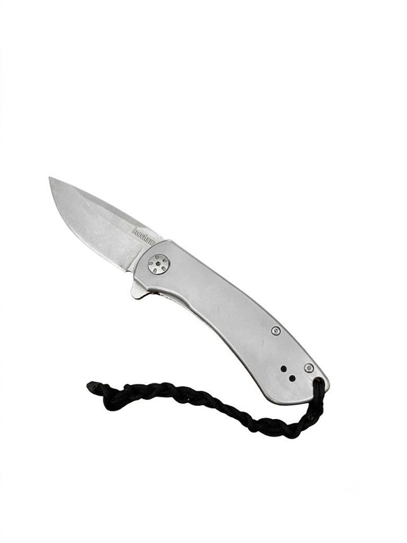Op de afbeelding: Een zilverkleurig inklapbaar zakmes met een metalen handvat en een scherp lemmet waarop de merknaam Kershaw is gegraveerd. Het mes is voorzien van een zwart gevlochten koord dat aan het uiteinde van het handvat is bevestigd voor extra grip.