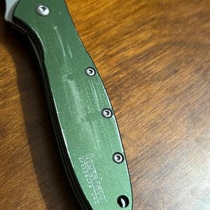 May include: Close-up of a green Kershaw SpeedSafe USA folding knife. The handle is a textured green with three visible screws. The blade is silver and partially visible. The knife is angled on a wooden surface.