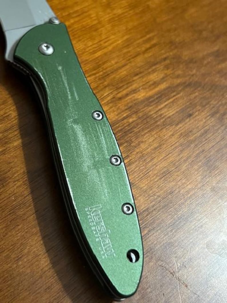 May include: Close-up of a green Kershaw SpeedSafe USA folding knife. The handle is a textured green with three visible screws. The blade is silver and partially visible. The knife is angled on a wooden surface.