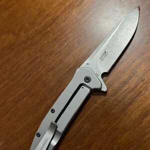 May include: A silver folding knife with a partially serrated blade. The knife has a pocket clip and the brand name "Kershaw" is visible. The blade has the text "2044 CAI SC13MOV". The knife is lying on a wooden surface.