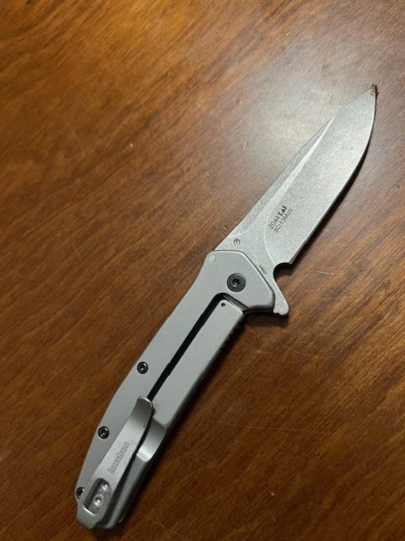May include: A silver folding knife with a partially serrated blade. The knife has a pocket clip and the brand name "Kershaw" is visible. The blade has the text "2044 CAI SC13MOV". The knife is lying on a wooden surface.