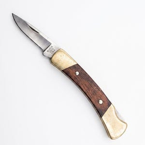 May include: A folding pocket knife with a silver-toned steel blade, a dark brown wood handle, and brass bolsters. The blade is stamped with the text BUCK 501A U.S.A. The knife features a curved handle design with two visible silver rivets.