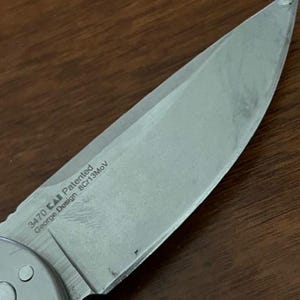 Op de afbeelding: Een zilverkleurig metalen lemmet van een zakmes met een puntige top en een scherpe snede. Het lemmet bevat gegraveerde tekst met 3470, een logo, Patented, George Design en 8Cr13MoV. Het metalen oppervlak heeft een geborstelde afwerking met lichte krassen.