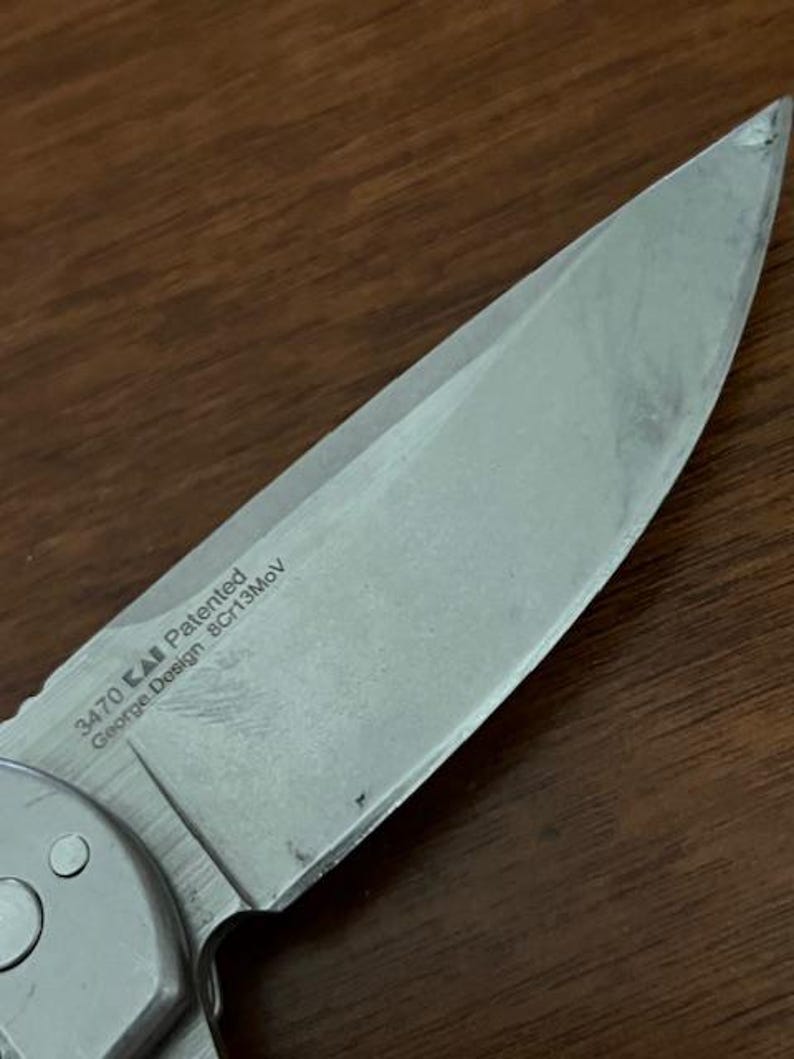 Op de afbeelding: Een zilverkleurig metalen lemmet van een zakmes met een puntige top en een scherpe snede. Het lemmet bevat gegraveerde tekst met 3470, een logo, Patented, George Design en 8Cr13MoV. Het metalen oppervlak heeft een geborstelde afwerking met lichte krassen.