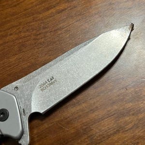 May include: Close-up of a silver folding knife with a partially visible handle. The blade has the text "2044 KAI 8Cr13MoV" printed on it. The knife is on a wooden surface.