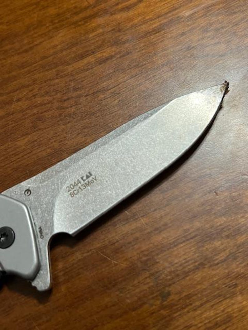 May include: Close-up of a silver folding knife with a partially visible handle. The blade has the text "2044 KAI 8Cr13MoV" printed on it. The knife is on a wooden surface.