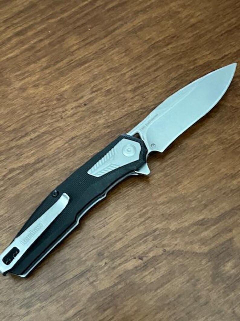 May include: A silver and black folding knife with a partially serrated blade. The knife has a black handle with a silver pocket clip and the brand name "Kershaw" printed on it. The blade is approximately 3 inches long.