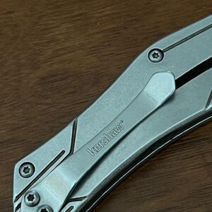 May include: Close-up of a silver Kershaw pocket knife with a pocket clip. The knife has a textured metal handle with geometric design elements and the brand name "Kershaw" is visible on the clip.