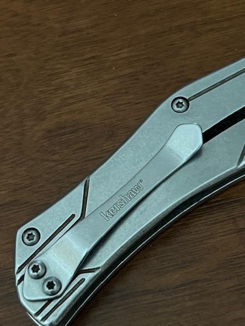 May include: Close-up of a silver Kershaw pocket knife with a pocket clip. The knife has a textured metal handle with geometric design elements and the brand name "Kershaw" is visible on the clip.