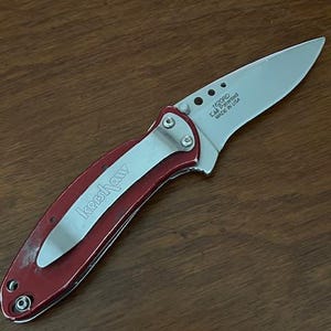 May include: A folding pocket knife with a metallic red handle and a silver pocket clip featuring the Kershaw brand name. The silver blade is partially extended and displays the text 1620RD, Cal Patented, and MADE IN USA near the pivot point.