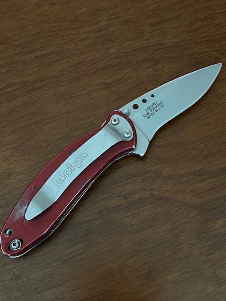 May include: A folding pocket knife with a metallic red handle and a silver pocket clip featuring the Kershaw brand name. The silver blade is partially extended and displays the text 1620RD, Cal Patented, and MADE IN USA near the pivot point.