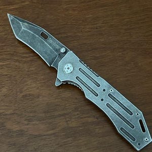 Puede incluir: Una navaja plegable con un acabado de metal gris desgastado. La hoja presenta una punta distintiva y la palabra Kershaw est&aacute; grabada cerca del pivote. El mango tiene una serie de cortes paralelos y tornillos expuestos de dise&ntilde;o utilitario.