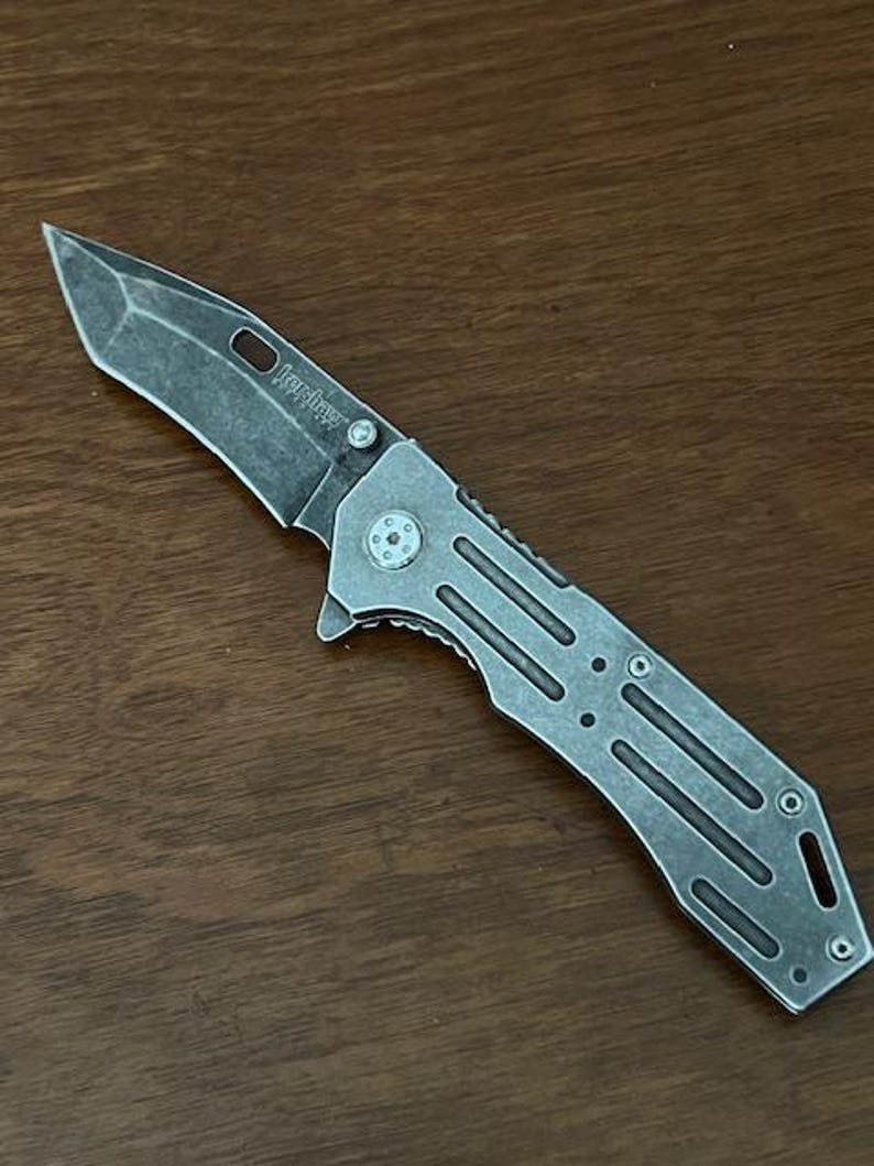 Puede incluir: Una navaja plegable con un acabado de metal gris desgastado. La hoja presenta una punta distintiva y la palabra Kershaw est&aacute; grabada cerca del pivote. El mango tiene una serie de cortes paralelos y tornillos expuestos de dise&ntilde;o utilitario.