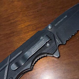 May include: Black folding knife with a serrated blade. The knife features a pocket clip with the "Kershaw" logo and the text "2021PROMO1 CAB" on the blade. The handle has a textured grip for secure handling.