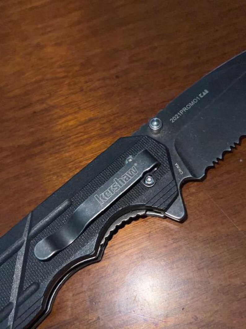 May include: Black folding knife with a serrated blade. The knife features a pocket clip with the "Kershaw" logo and the text "2021PROMO1 CAB" on the blade. The handle has a textured grip for secure handling.