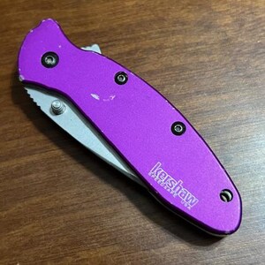 May include: A purple folding knife with a silver blade. The knife has a textured handle and the brand name "Kershaw" is printed on the side. The knife is closed and resting on a wooden surface.