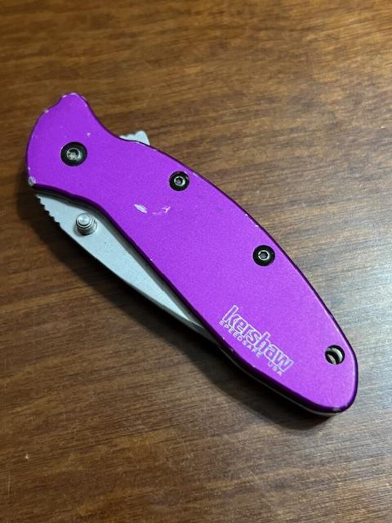 May include: A purple folding knife with a silver blade. The knife has a textured handle and the brand name "Kershaw" is printed on the side. The knife is closed and resting on a wooden surface.