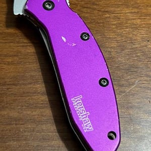 May include: A purple folding knife with a silver blade. The knife handle has three black screws and the brand name "Kershaw" printed in white. The knife is on a wooden surface.