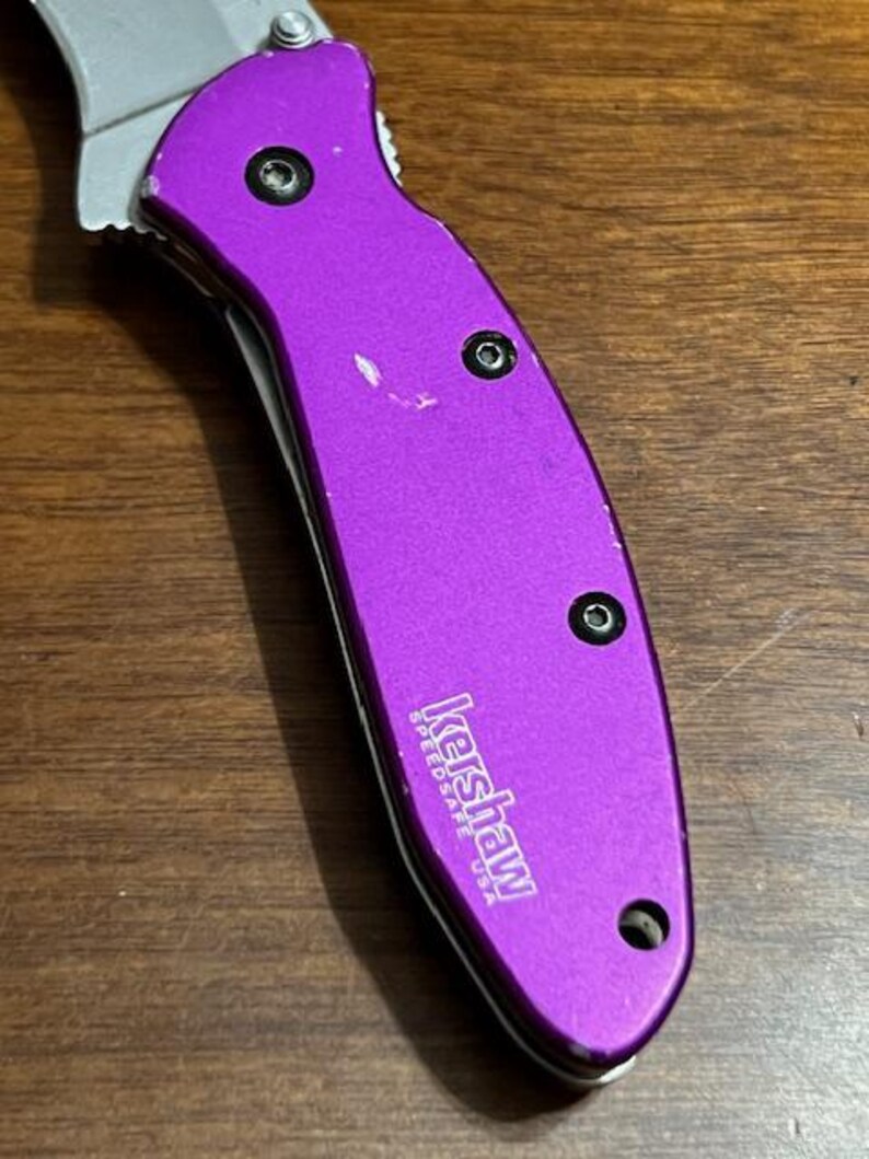 May include: A purple folding knife with a silver blade. The knife handle has three black screws and the brand name "Kershaw" printed in white. The knife is on a wooden surface.