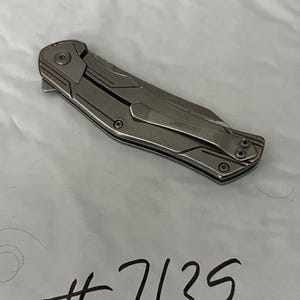May include: A silver folding knife with a pocket clip. The knife has a textured handle and a blade that folds into the handle. The text "Kershaw" is visible on the clip. The number "7135" is handwritten in black ink.