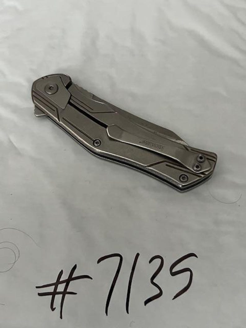 May include: A silver folding knife with a pocket clip. The knife has a textured handle and a blade that folds into the handle. The text "Kershaw" is visible on the clip. The number "7135" is handwritten in black ink.