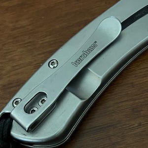 Op de afbeelding: Een zilverkleurig metalen zakmeshandvat met een zakclip waarop de merknaam Kershaw is gegraveerd. Het handvat is bevestigd met drie kleine stervormige schroeven en bevat een zwart koordje dat aan de onderkant is bevestigd.