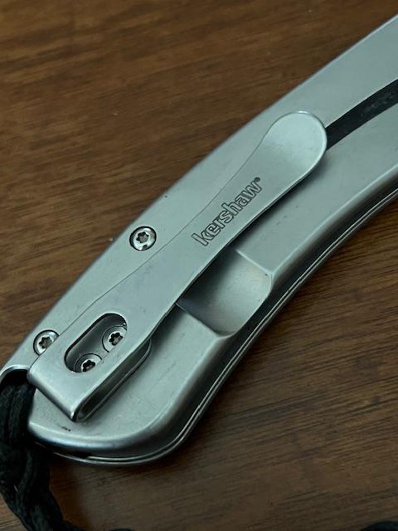 Op de afbeelding: Een zilverkleurig metalen zakmeshandvat met een zakclip waarop de merknaam Kershaw is gegraveerd. Het handvat is bevestigd met drie kleine stervormige schroeven en bevat een zwart koordje dat aan de onderkant is bevestigd.
