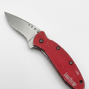 May include: A folding pocket knife with a metallic silver blade and a red handle. The handle features the text Kershaw SpeedSafe USA and the number 1660. The blade has three circular cutouts near the base, and the handle is secured with black screws.