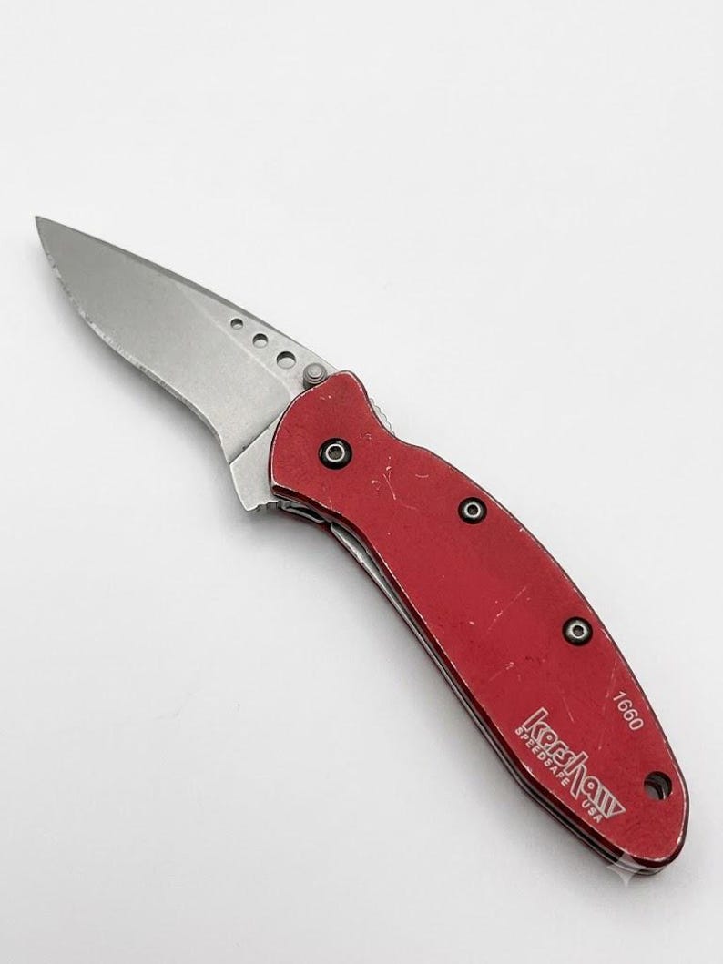 May include: A folding pocket knife with a metallic silver blade and a red handle. The handle features the text Kershaw SpeedSafe USA and the number 1660. The blade has three circular cutouts near the base, and the handle is secured with black screws.