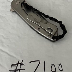 Op de afbeelding: Een zilverkleurig inklapbaar zakmes met een metalen handvat, een zakclip met de merknaam Kershaw en een zwart gevlochten koord aan het uiteinde. Het nummer #7100 is met zwarte inkt onder het mes met de hand geschreven.