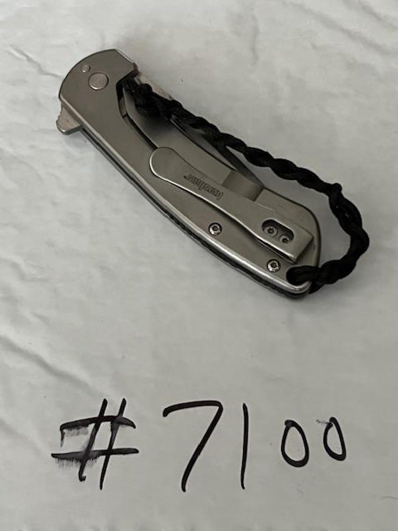 Op de afbeelding: Een zilverkleurig inklapbaar zakmes met een metalen handvat, een zakclip met de merknaam Kershaw en een zwart gevlochten koord aan het uiteinde. Het nummer #7100 is met zwarte inkt onder het mes met de hand geschreven.