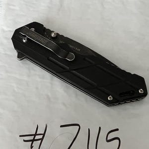 May include: Black folding knife with a pocket clip and the brand name "Kershaw". The knife has a textured handle and a blade. The number "7115" is written in black marker below the knife.