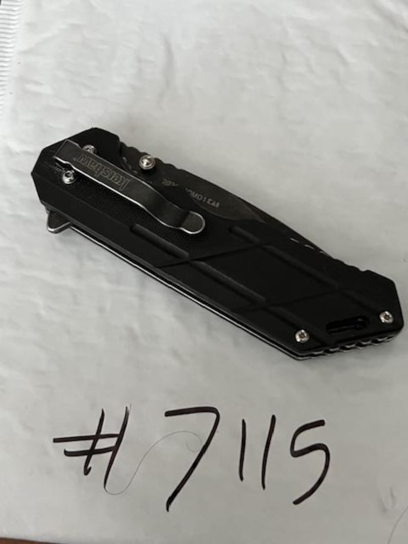 May include: Black folding knife with a pocket clip and the brand name "Kershaw". The knife has a textured handle and a blade. The number "7115" is written in black marker below the knife.